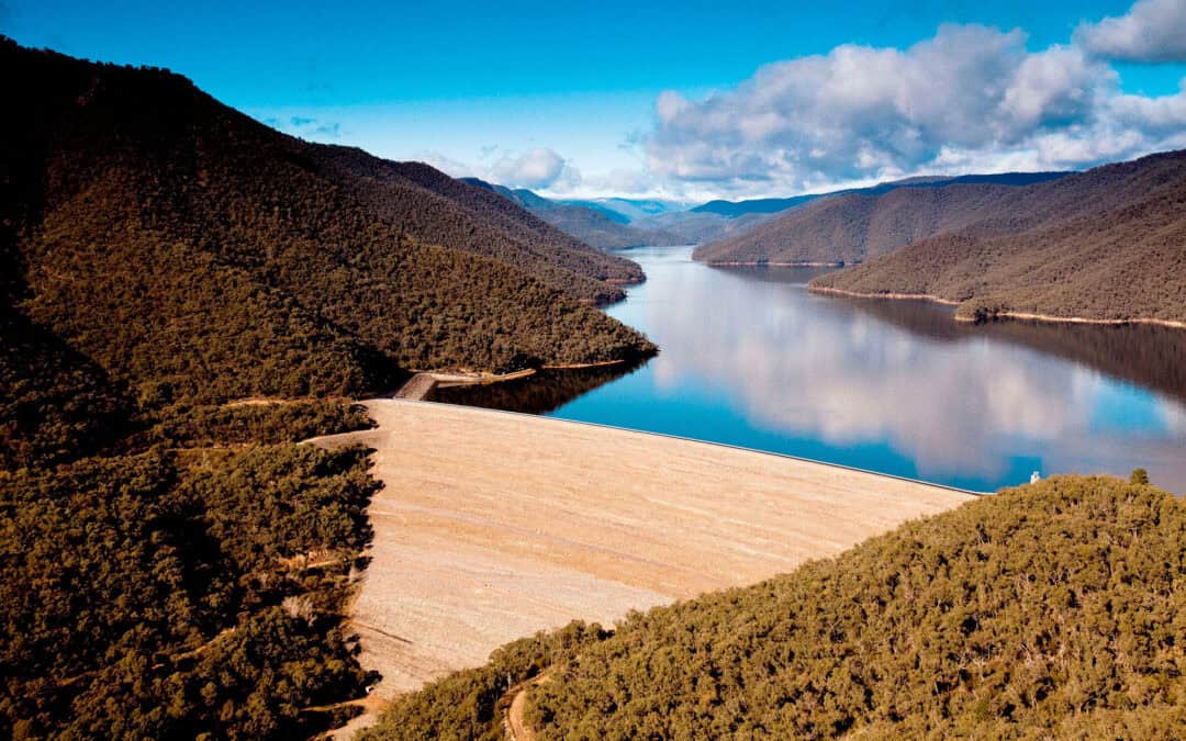 Reservoir dam in a mountain valley surrounded by forested hills beneath a blue sky.