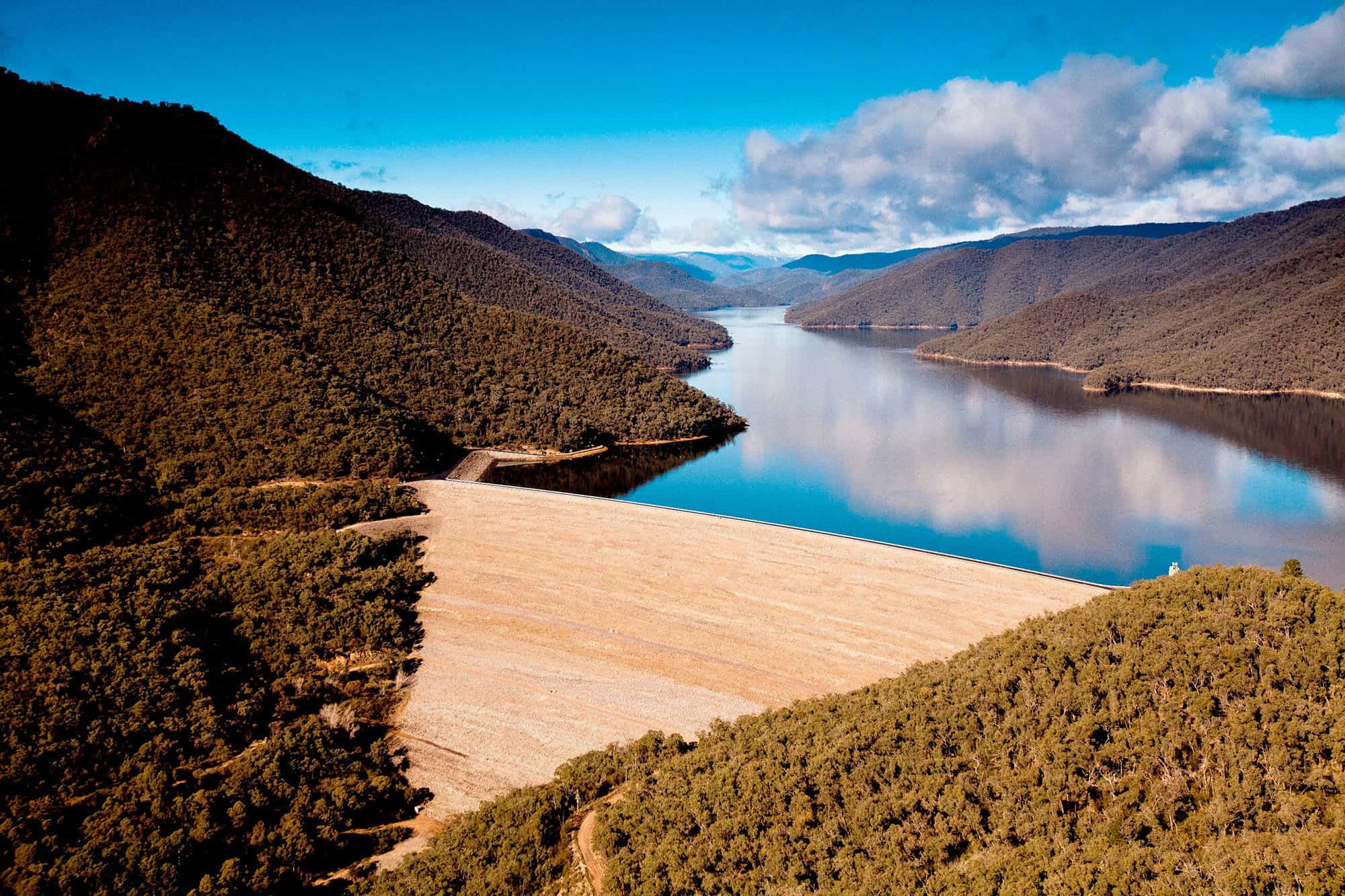 Reservoir dam in a mountain valley surrounded by forested hills beneath a blue sky.